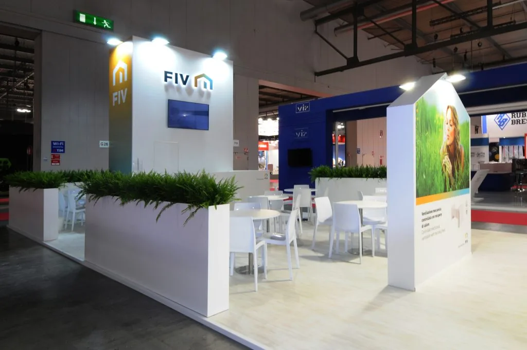 innovazione per sanità e industria Booth espositivo moderno con arredi bianchi, piante verdi e pannelli promozionali, allestito in una fiera.