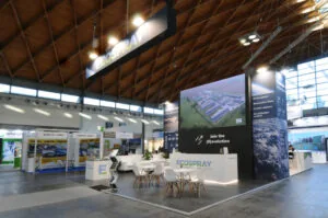 Stand ECOSPRAY in fiera, con display verticale e slogan ambientale, sedute bianche, schermi digitali e grafica dedicata a green power, carburanti sostenibili e cattura del carbonio.
