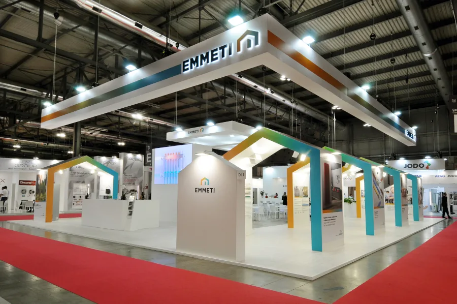 Stand EMMETI in fiera, con design moderno e strutture geometriche colorate, sedute bianche, schermi digitali e branding centrale su sfondo bianco.