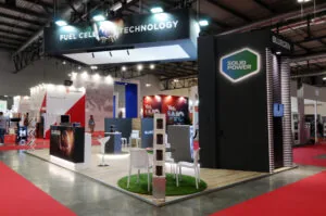 Stand BLUEGEN di SOLID POWER in fiera, dedicato alla tecnologia delle celle a combustibile, con design moderno, sedute bianche, schermi informativi e dettagli in nero, bianco e rosso su pedana in legno.